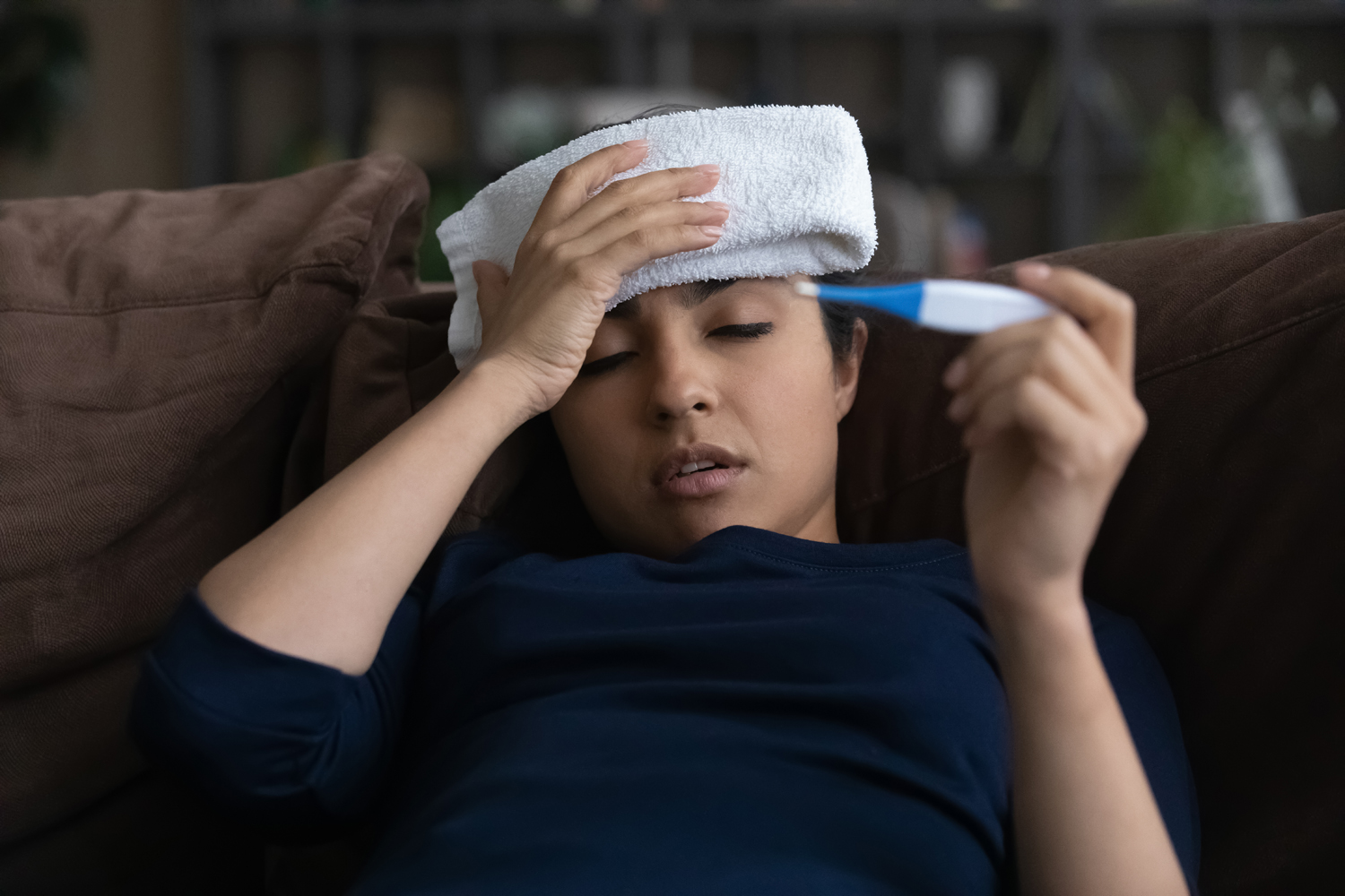 A sick woman slouches on the sofa. She holds a folded hand towel over her forehead while checking the temperature reading on her thermometer.
